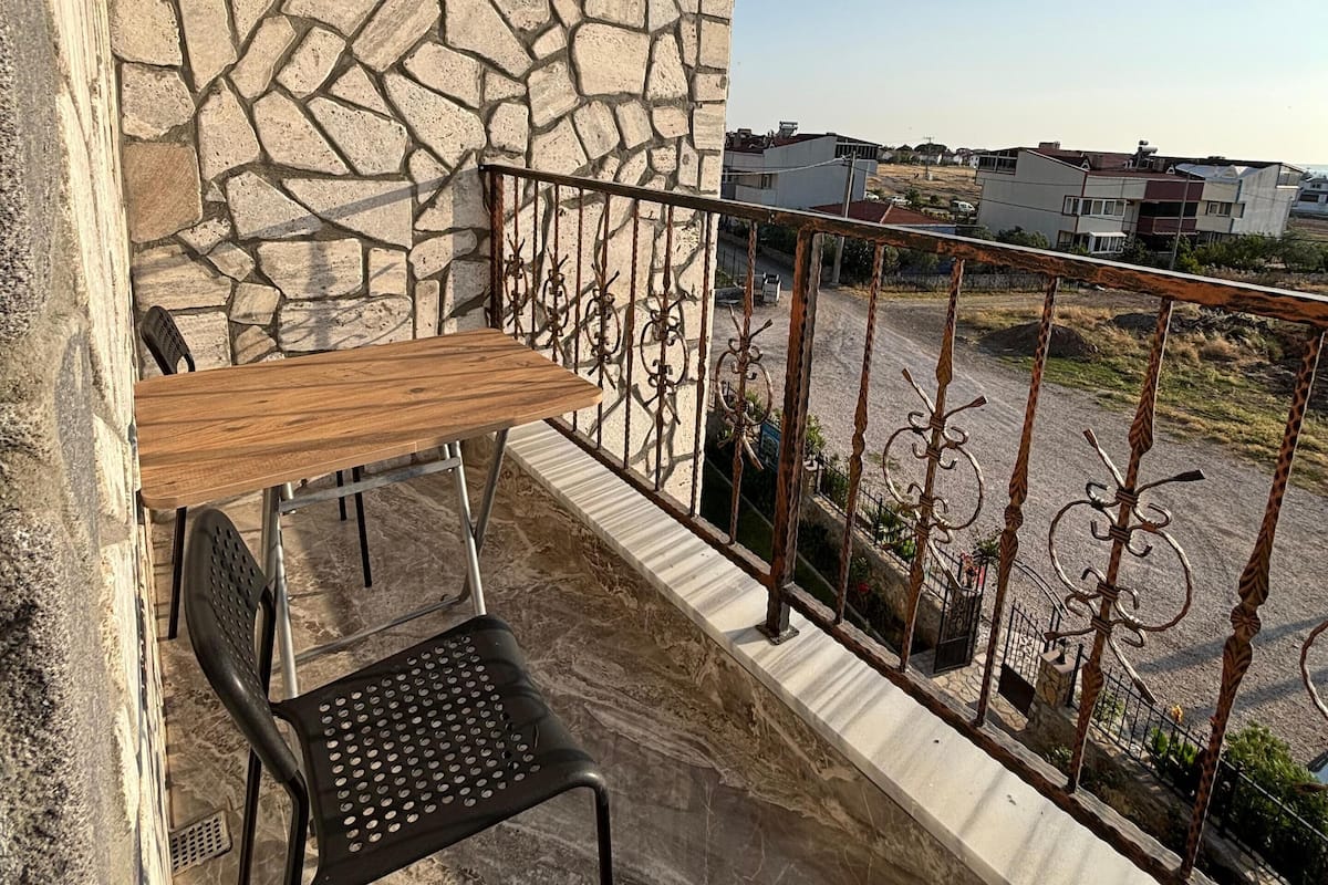 Un balcón con pared de piedra, una mesa de madera, una silla negra y una barandilla de metal con vista a un estacionamiento.