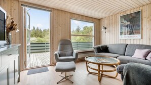 Ein modernes Wohnzimmer mit einem grauen Sofa, einem grauen Sessel und einem kleinen runden Tisch. Ein großes Fenster bietet Blick auf Bäume und eine Terrasse.