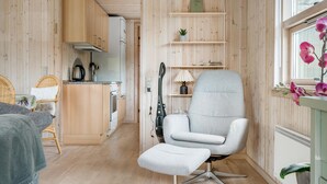 Ein gemütliches Zimmer mit grauem Sessel, einem kleinen Hocker, Holzkästen, einer Küchenzeile und einem Fenster mit Blick.