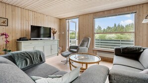 Ein gemütliches Wohnzimmer mit Fernseher, grauen Sofas, einem runden Couchtisch und einem großen Fenster mit Blick auf Bäume.