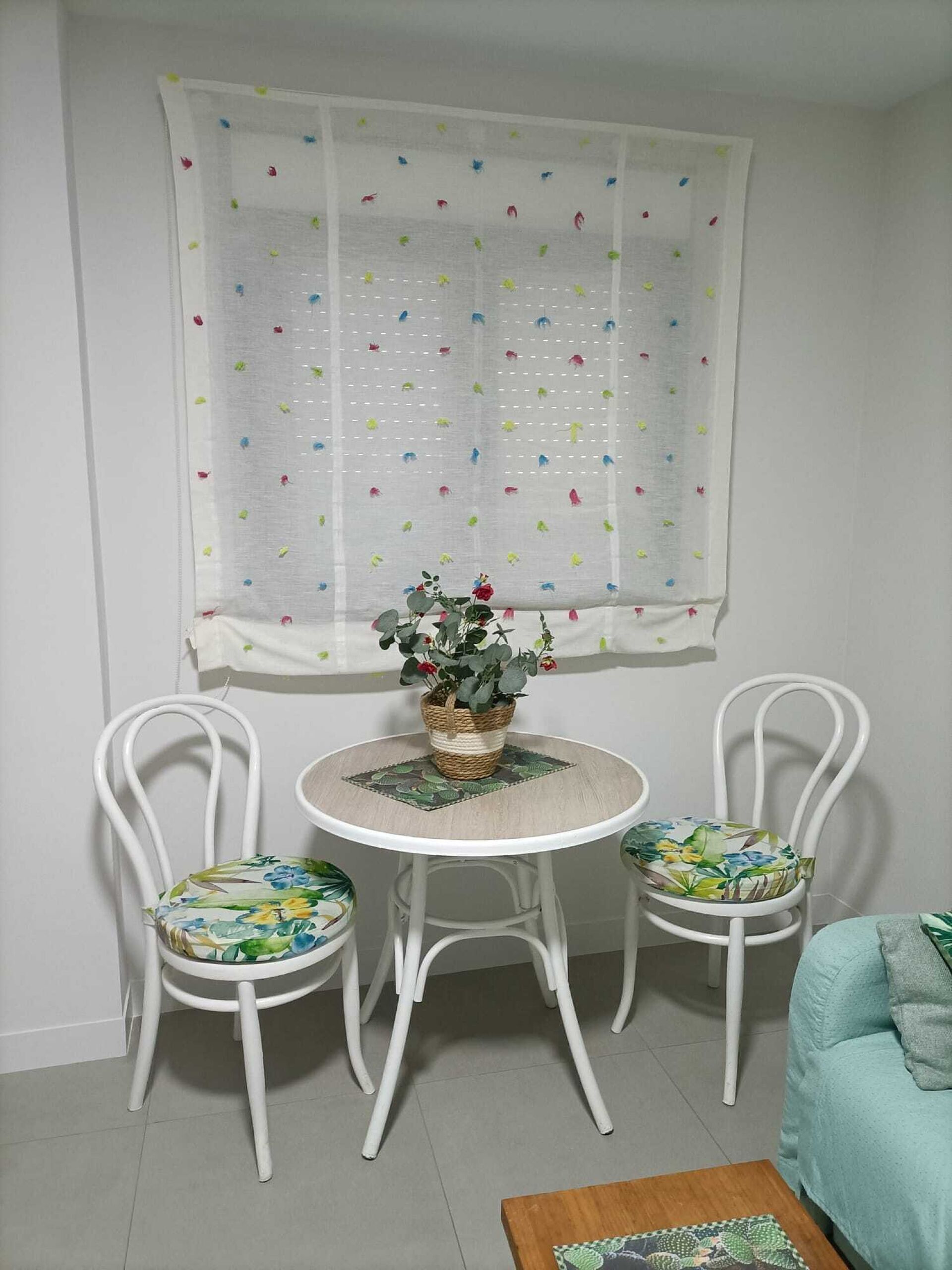 Ein kleiner Essbereich mit rundem Tisch und zwei Stühlen, einem Fenster mit leichtem Vorhang und einem Blumentopf auf dem Tisch.