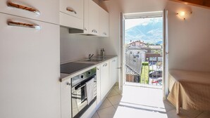 Eine moderne Küche mit weißen Schränken, einer Edelstahlspüle und einem Backofen. Ein Balkon mit Blick auf Häuser und Berge ist sichtbar.