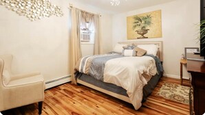 Un dormitorio con cama, una silla, una araña, un cuadro y una ventana con cortinas.