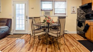 Un comedor con mesa de vidrio y sillas acolchadas, una puerta, y una cocina con alacenas de madera y estufa.