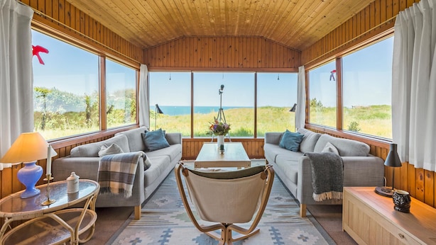 A cozy room with wooden walls and large windows offering a view of the sea. There is a sofa, a chair, and a small table with a lamp. - Lovely home in Hundested with WiFi (Hundested)
