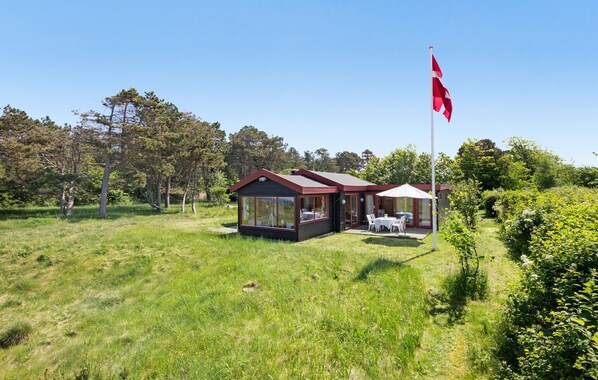 Une cabane dotée d’une paroi vitrée, un espace repas avec des sièges à l’extérieur et un mât avec un drapeau.