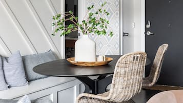Ein modernes Wohnzimmer mit einem dunklen Holztisch, Korbstühlen, einer weißen Vase mit Grünpflanzen und ornamentalem Wandpapier.
