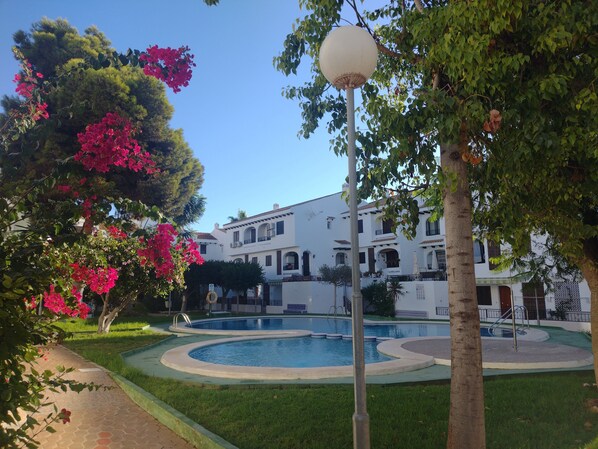 A pool area with a white building in the background, surrounded by greenery and pink flowers.