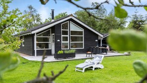 Et moderne, sort hus med store vinduer, en terrasse med bålsted og en græsplæne med en liggestol.