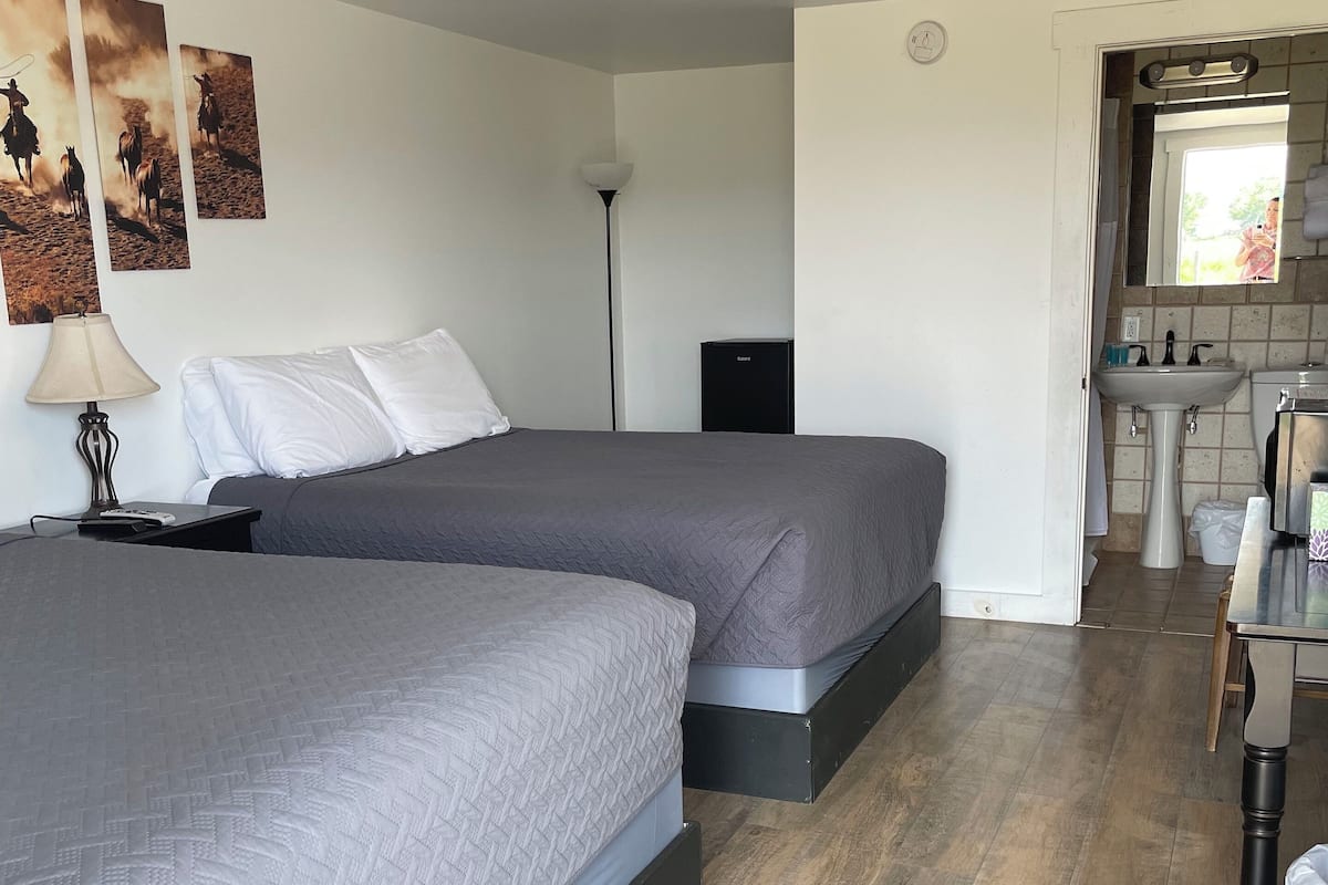 A hotel room with two beds, a desk, a lamp, a small black speaker, and a bathroom visible through an open door.