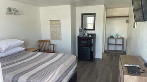 Un dormitorio con una cama, una silla de madera, un escritorio pequeño con un microondas y un televisor montado en la pared.