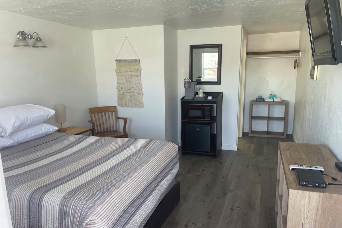 A bedroom with a bed, a wooden chair, a small desk with a microwave, and a TV mounted on the wall.