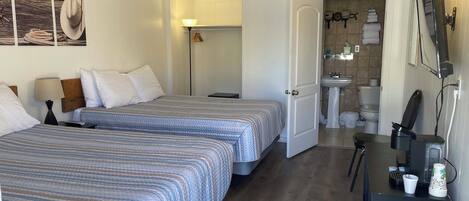 A hotel room with two beds, a desk, a chair, a trash bin, a bathroom with a sink and toilet, and a wall with a cowboy hat and rope.
