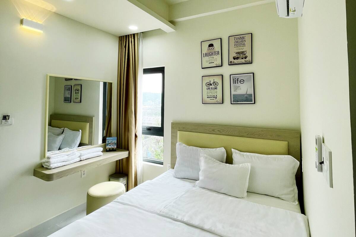 A hotel room with a large bed, a bedside table, a mirror, and a window with curtains. There are wall-mounted pictures and an air conditioning unit.
