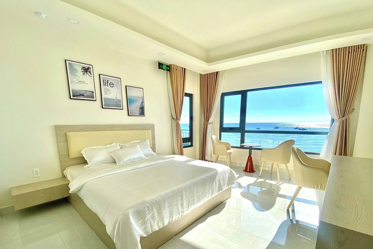 A modern hotel room with a large bed, a seating area with chairs, and a view of the sea.