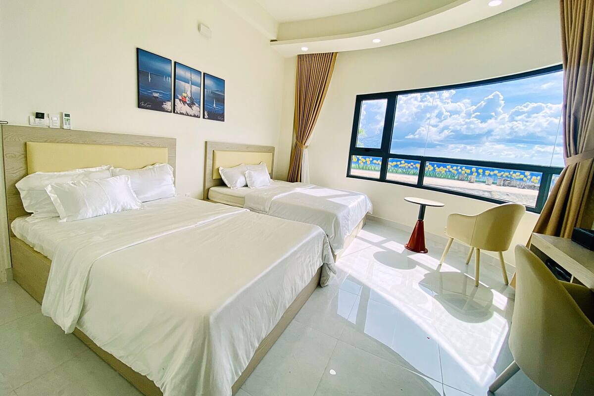A hotel room with two beds, a small table, and a chair. There are paintings on the wall and a large window with a view.