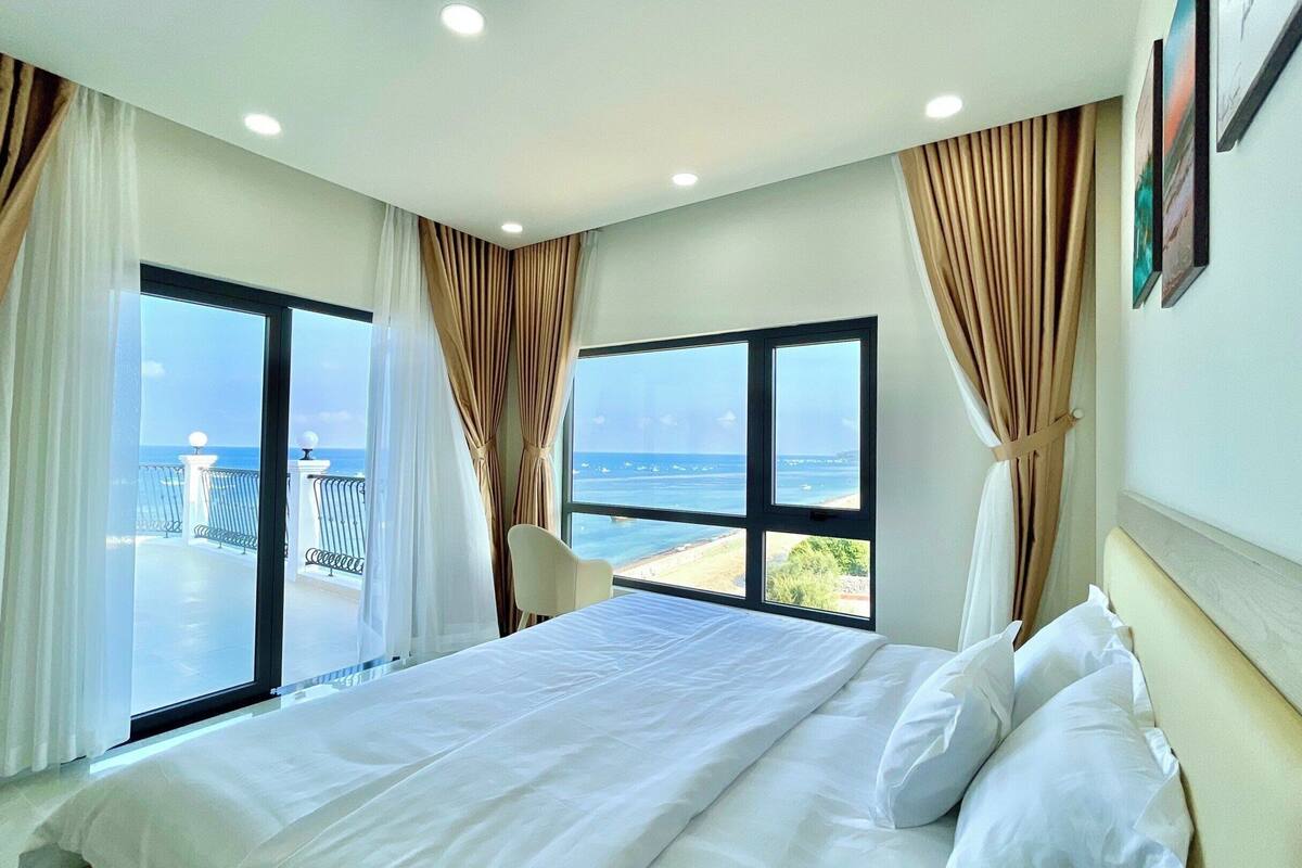 A hotel room with a large bed, two windows with curtains, a view of the sea, and a balcony with a railing.