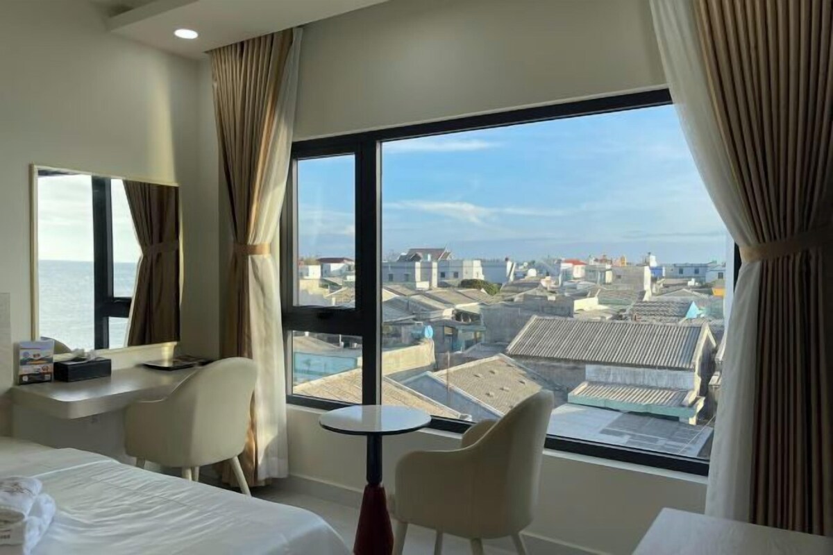 A hotel room with a bed, two chairs, a small table, a mirror, and a large window offering a city view.