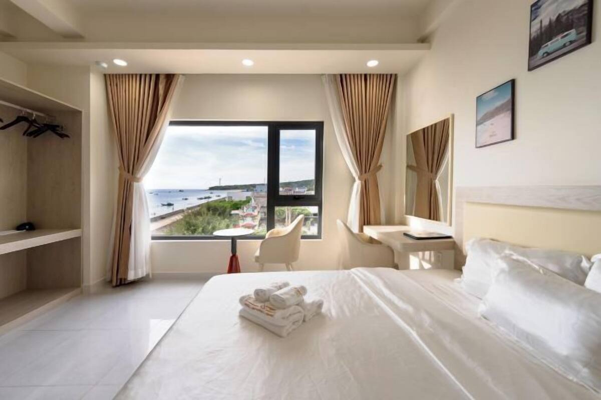 A hotel room with a large bed, a desk, a chair, a view of the sea, and a wardrobe.