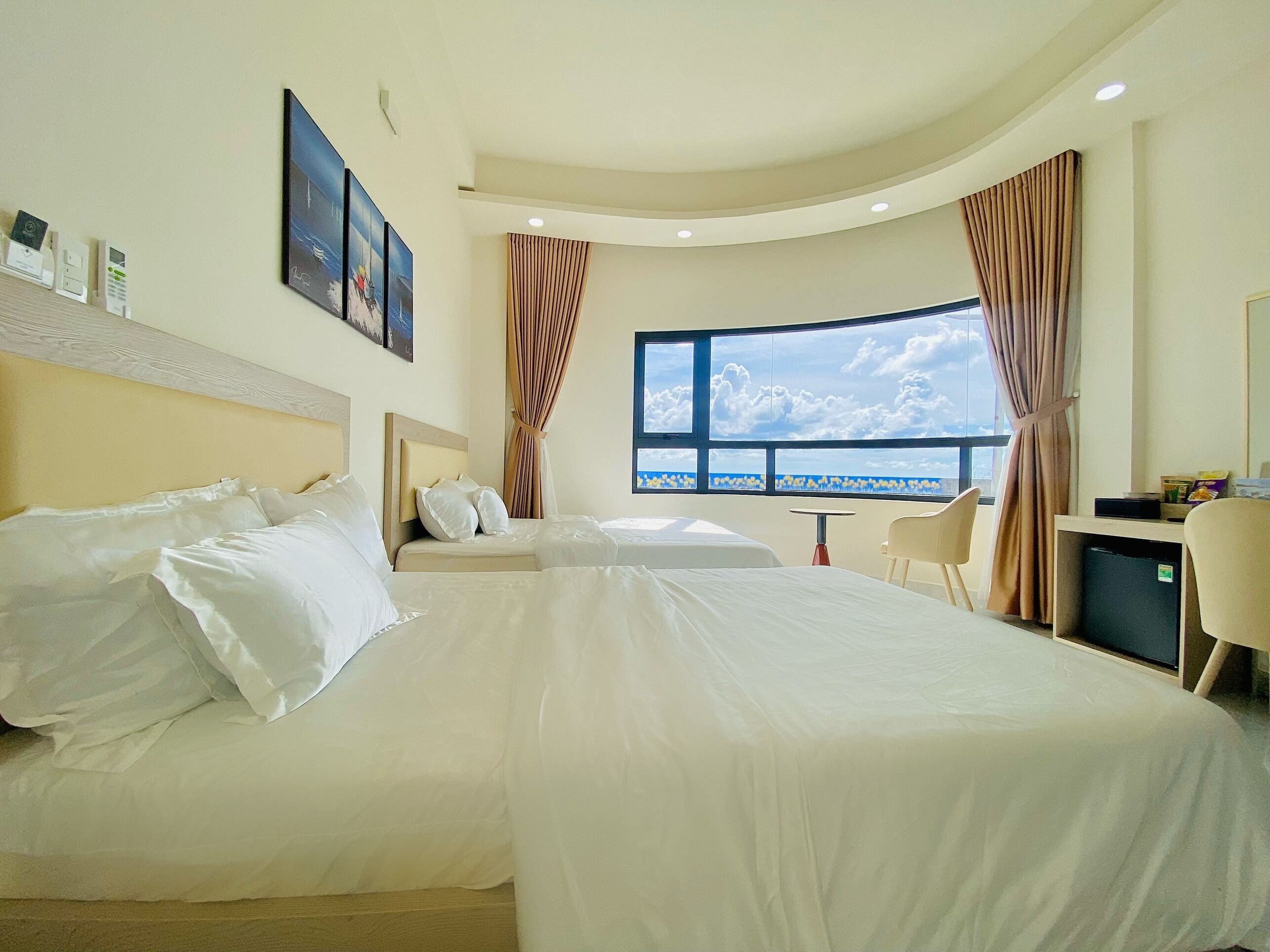 A hotel room with two beds, a desk, a chair, a small table, and a large window with a view of the sea.