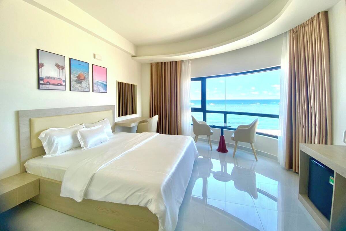 A hotel room with a bed, a desk, two chairs, a TV, and a large window with a view of the ocean.