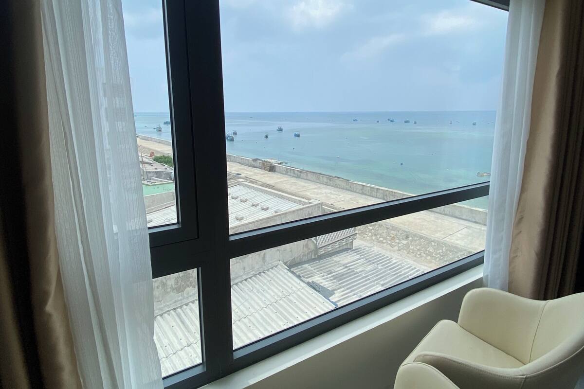 A room with a large window overlooking a beach and the sea. There are two white chairs in the room.