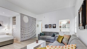 A modern living room with a grey sofa, a white coffee table, and a grey bed. There is a bookshelf with books and framed pictures on the wall. - Cozy Studio with private Outdoor Space - FromHometoRome (Roma)