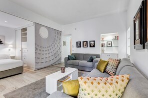 Un salon moderne avec un canapé gris, une table basse blanche et un lit gris. On y trouve une bibliothèque remplie de livres et des tableaux encadrés accrochés au mur.