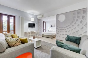 Un salon moderne avec un canapé, une table basse et un téléviseur. Il y a également un coin repas avec des chaises et une table. La pièce est dotée d’une grande fenêtre et d’un mur orné d’un motif circulaire.