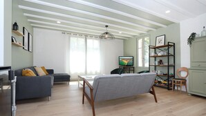 A modern living room with a grey sofa, a wooden coffee table, and a TV on a stand. There is a bookshelf with books and decorative items, and a window with sheer curtains allowing natural light.