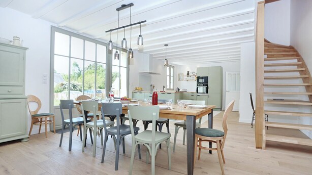 A spacious dining area with a wooden table and chairs, a kitchen in the background, and a staircase leading to an upper level.