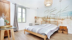 A bedroom with a wooden bed, a glass table with decorative items, a window with curtains, and a wall mural of tall grasses.