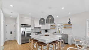 Una cocina moderna con armarios blancos, una isla grande con taburetes, electrodomésticos de acero inoxidable e iluminación colgante.
