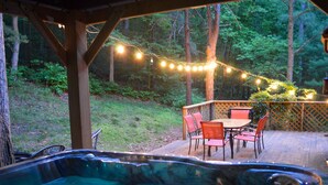 A hot tub with a wooden deck, surrounded by chairs and a table, with string lights and a wooden fence in a wooded area. - Cabin in the Trees, Hot Tub, Fire Pit (Rockbridge)