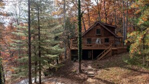 A rustic cabin surrounded by autumn foliage, with a wooden deck and a staircase leading to the entrance. - Cabin in the Trees, Hot Tub, Fire Pit (Rockbridge)
