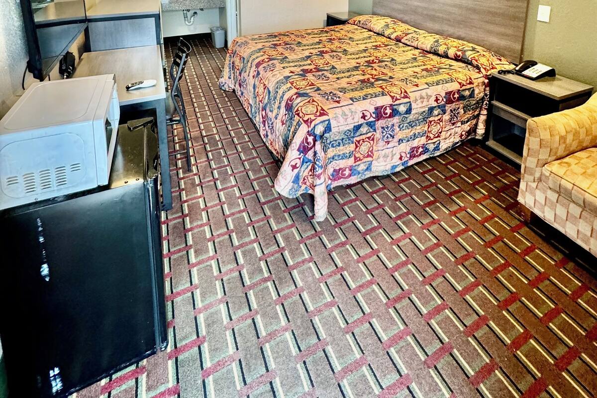 A hotel room with a large bed, a desk, a chair, a dresser, and a small table with a lamp.