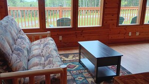 Interior acogedor de una cabaña con sofá de madera, una mesita y un tapete. Amplias ventanas ofrecen vistas a una terraza de madera y follaje.