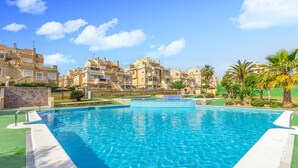 A swimming pool with a clear blue water, surrounded by a green deck and a stone wall. In the background, there are multi-story buildings with balconies and a clear sky. - 2 bedroom stunning apartment in Torrevieja (Torrevieja)