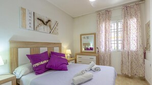 A bedroom with a wooden headboard, a bed with purple pillows, a nightstand with a lamp, a dresser, a mirror, and a window with floral curtains. - 2 bedroom stunning apartment in Torrevieja (Torrevieja)