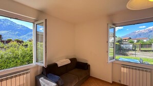 A small, cozy room with a single brown sofa, a small table, and two windows offering a mountain view. - Beautiful apartment in Domaso with WiFi (Domaso)