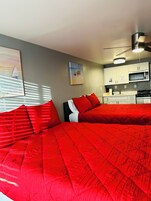 Un dormitorio con dos camas, ventilador de techo, un cuadro y una kitchenette.