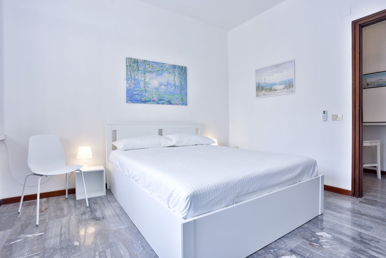 Un dormitorio con una cama blanca, una silla, una mesita de noche y dos cuadros en la pared.