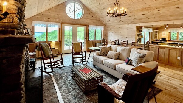 A spacious log cabin interior with a high ceiling, a stone fireplace, a cozy seating area with a sofa and chairs, a dining table, and a kitchen area.