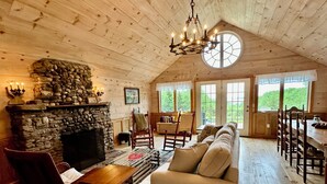 A cozy log cabin interior with a stone fireplace, wooden furniture, and large windows offering a view of the outdoors.