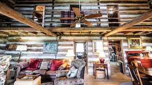 A rustic cabin interior with a wooden ceiling, a ceiling fan, a leather sofa, a patterned armchair, a dining table, and a red car in the background.
