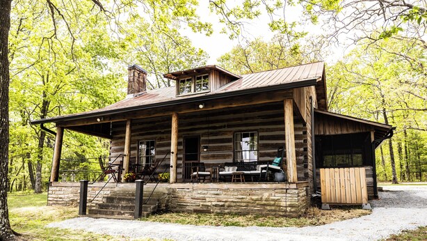 A rustic log cabin with a porch, stone foundation, and a chimney.
