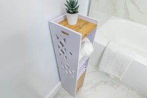 Een hoek van een badkamer met een witte badkuip, een witte handdoek aan de muur en een grijze opbergunit met een houten plank erboven.