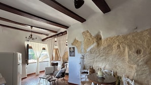 Quarto rústico com paredes de pedra aparente, teto de madeira e área de refeições com cadeiras de vime branco.