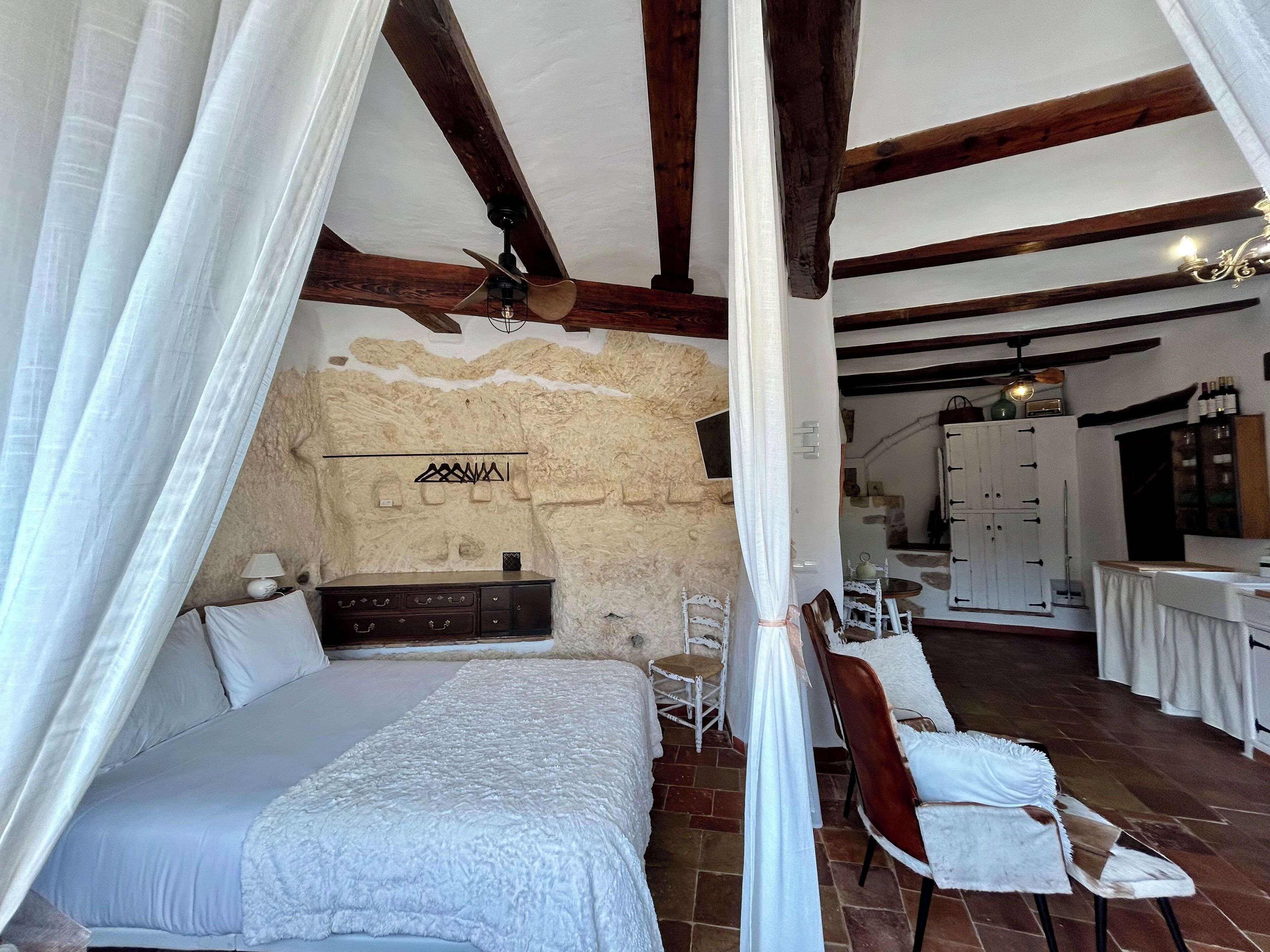A bedroom with a bed, a nightstand, a lamp, and a stone wall. There is a dining area with a table and chairs in the background.