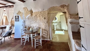 Um interior rústico com paredes de pedra aparente, uma área de jantar com mesa redonda e cadeiras brancas, e uma escada que leva a um andar superior.